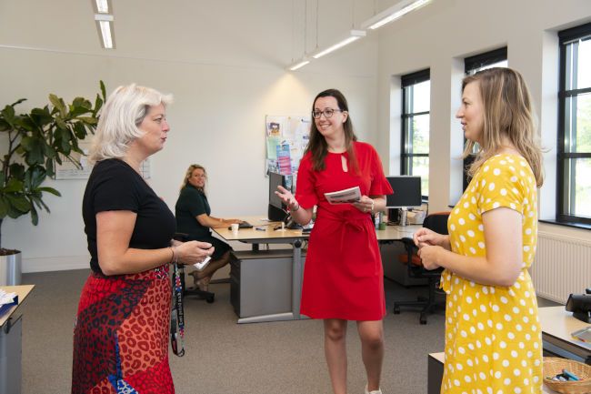 Drie vrouwelijke collega's staan in een open kantoorkamer en zijn prettig in overleg